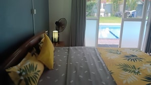 Quarto com cama, mesa de cabeceira, abajur, ventilador e vista para uma piscina.