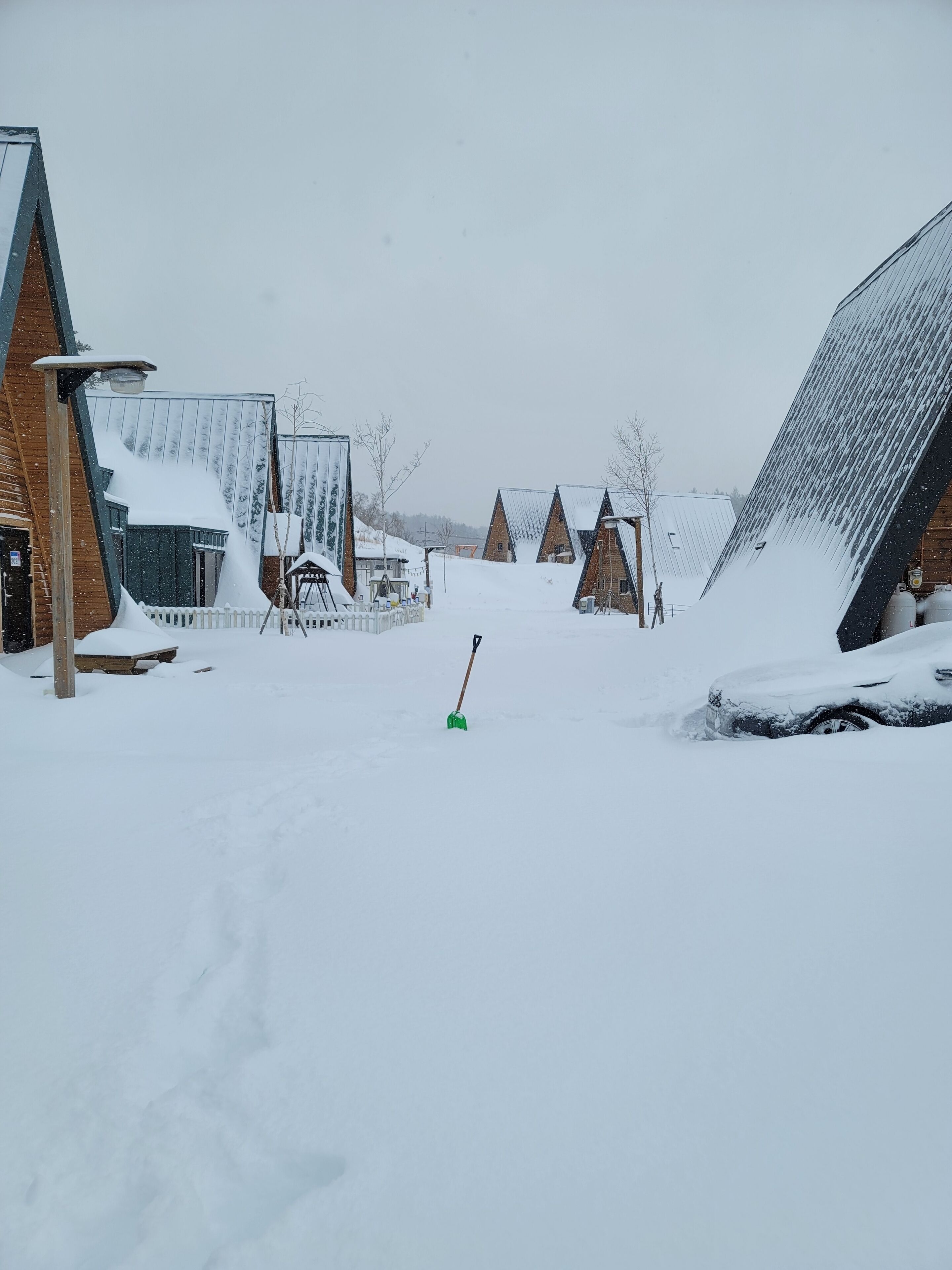 A snowy village with traditional wooden houses, a green shovel, and a car covered in snow.