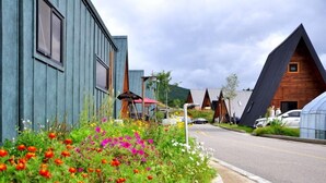 A street with modern buildings, one with a unique triangular roof and another with a sloped design. There are colorful flowers in the foreground and a clear sky.