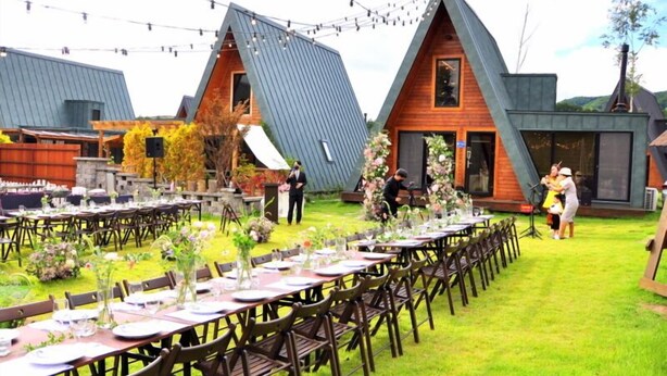 A wedding reception set up outdoors with multiple dining tables, guests, and a stage for speeches.