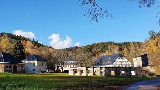 Un complejo histórico de edificios con varias estructuras, incluyendo un edificio central grande y edificios más pequeños adyacentes, situado en un paisaje rural con una colina boscosa al fondo.