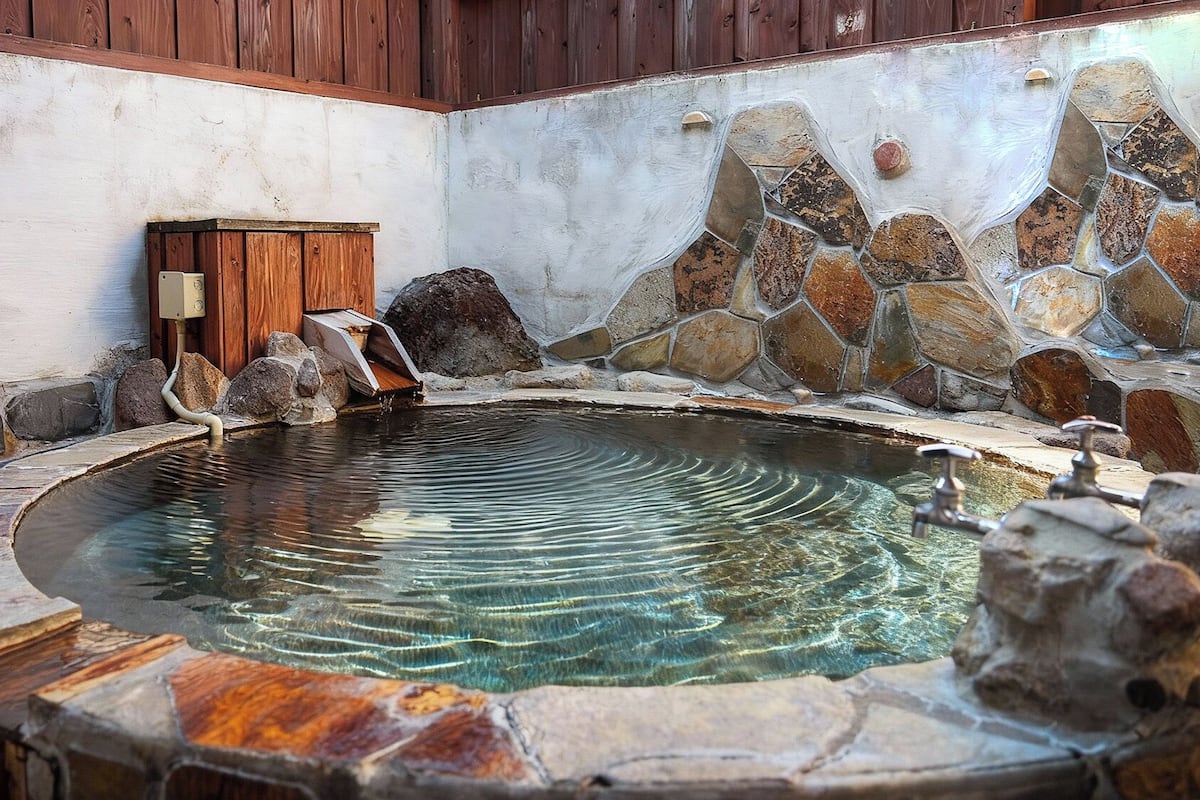 Uma área de águas termais com piscina de ladrilho, bancos de madeira e parede de pedra.