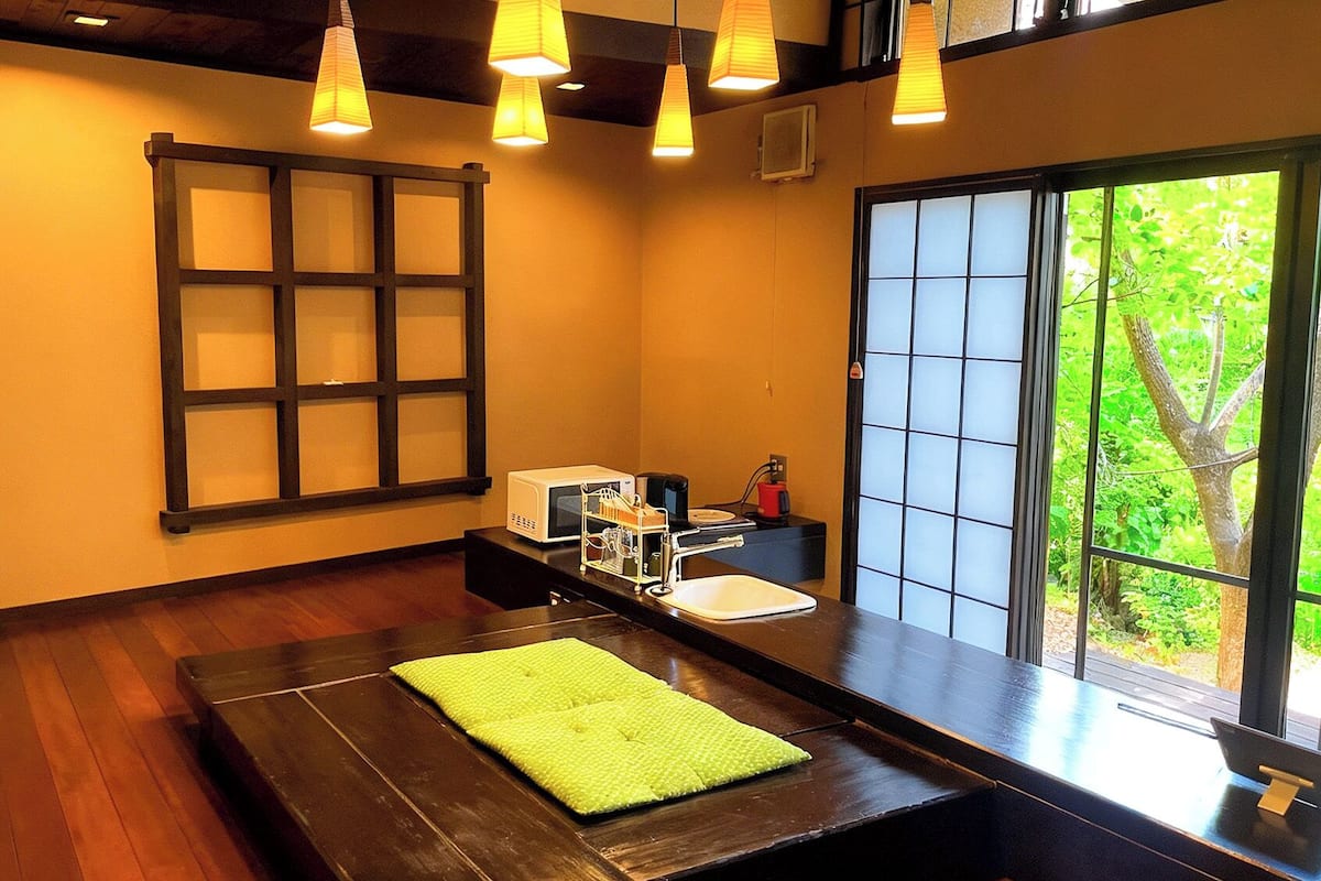 Quarto com piso de madeira, tapete verde, micro-ondas, cafeteira e janela com vista para vegetação.