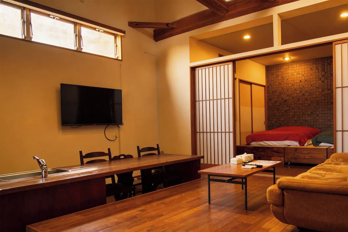 Espaço compacto com mesa de jantar de madeira, cadeiras, sofá, TV fixada na parede e porta deslizante que dá acesso a um quarto com cama e manta vermelha.