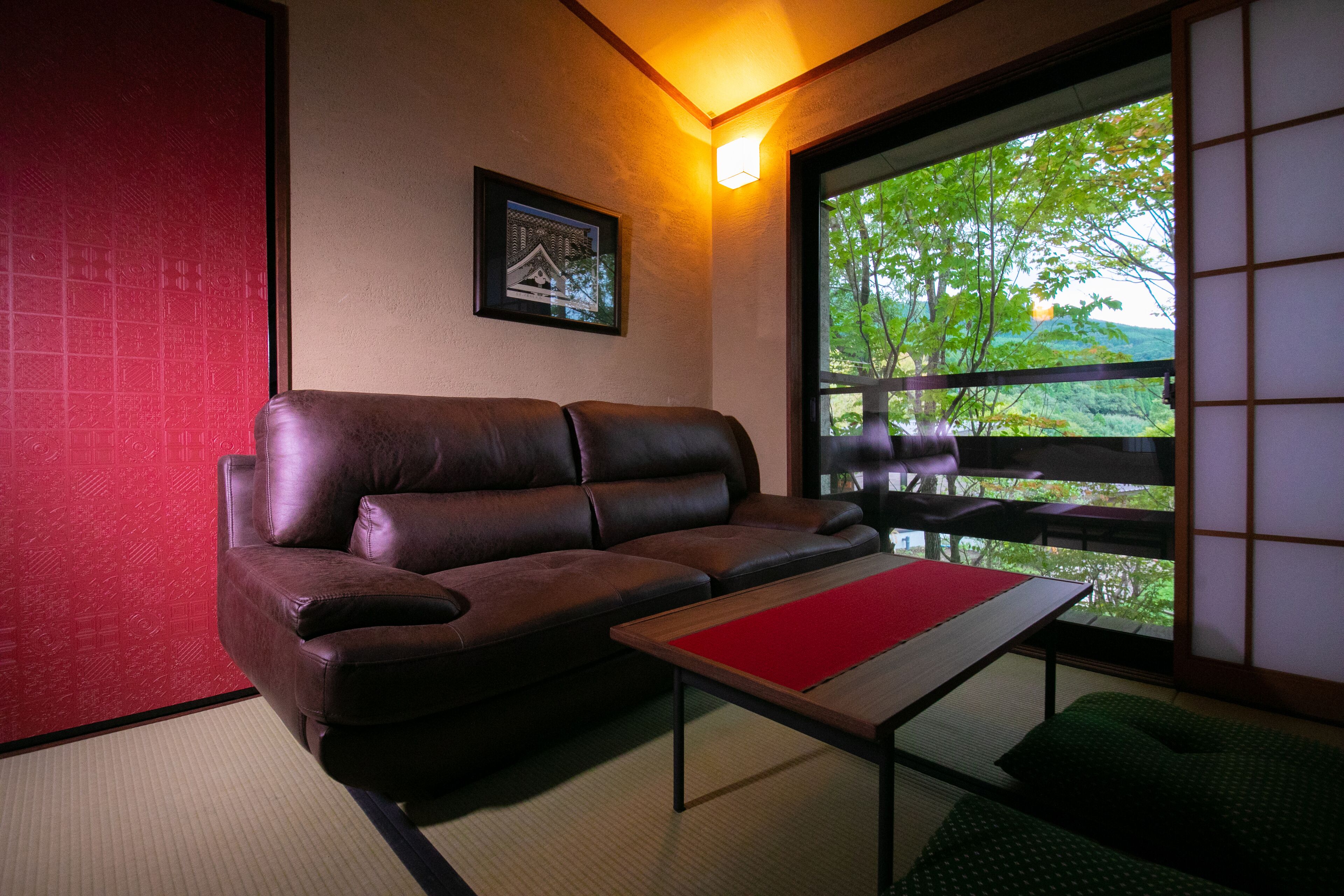Ein Zimmer mit einem braunen Ledersofa, einem kleinen Tisch mit einer roten Unterlage und einem großen Fenster mit Blick auf Grünflächen.