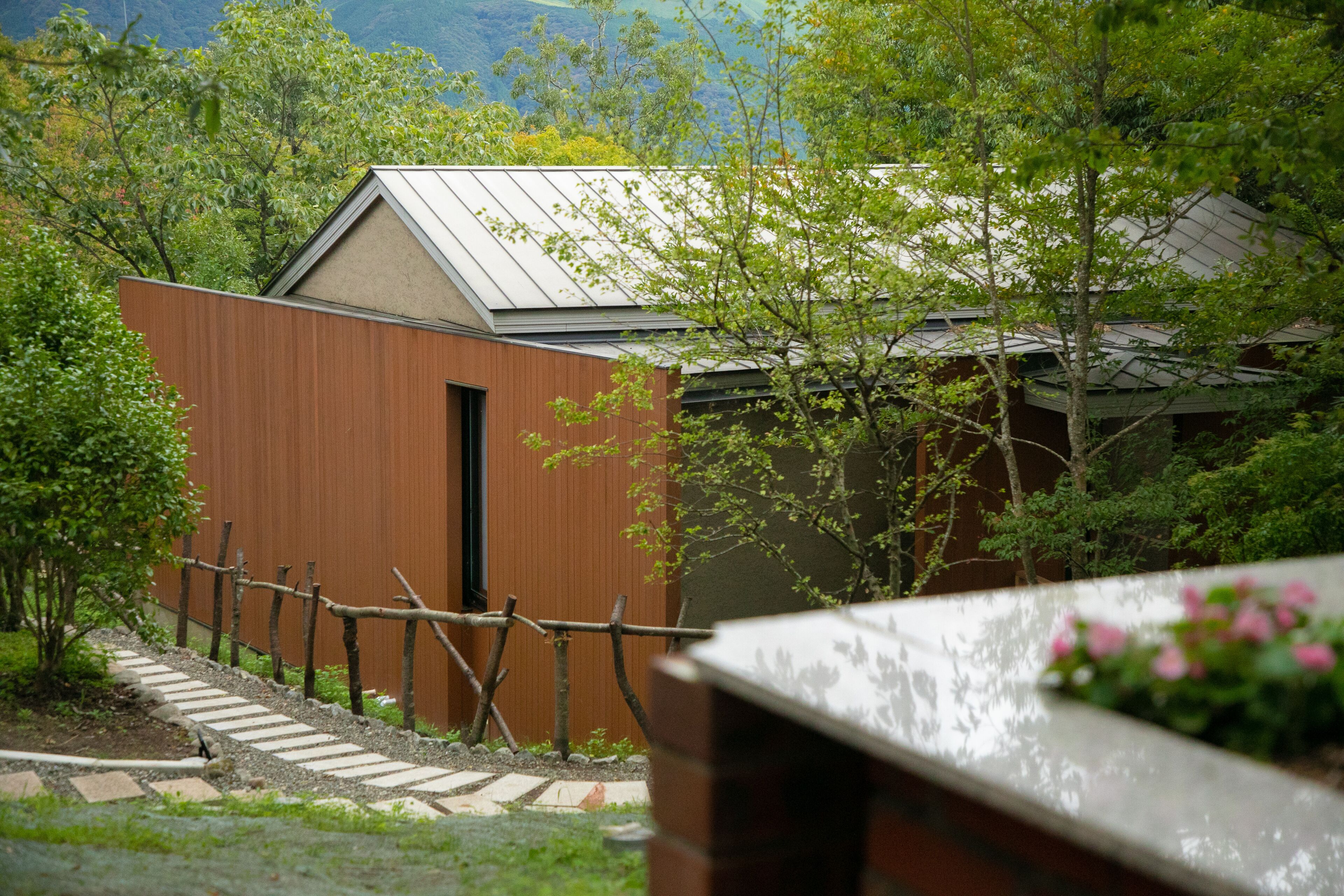 A modern building with a sloped roof, surrounded by lush greenery and a wooden fence.