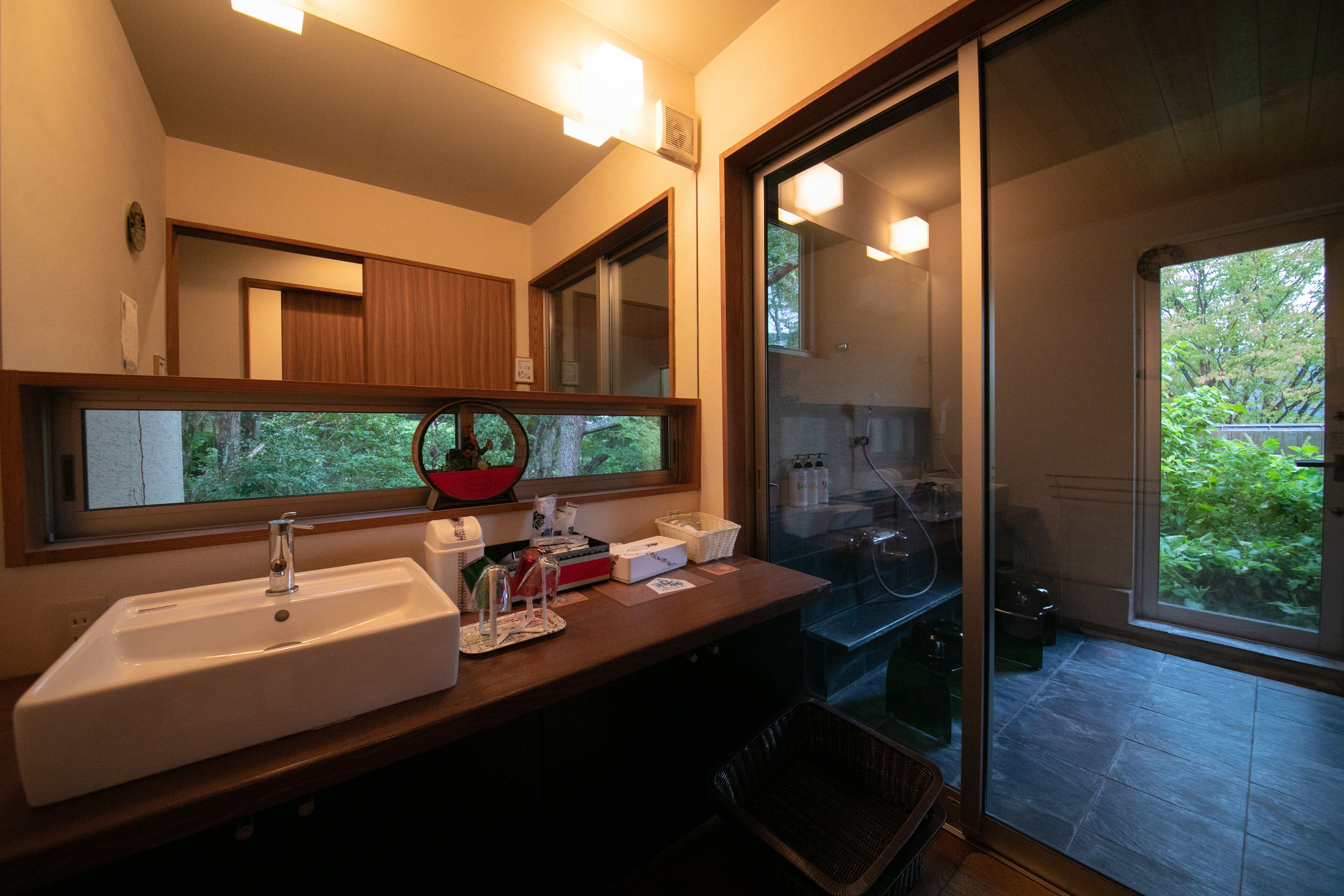 A bathroom with a large mirror, a sink, and a window overlooking greenery.