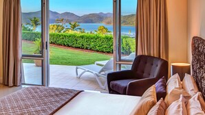 A hotel room with a large bed, a dark armchair, a small table, and a view of a landscaped garden and lake. - Bella Vista East 3 Luxury Ocean Views Large Flat Grassed Area + Buggy (Whitsundays)