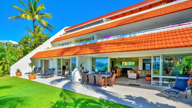 A modern two-story house with a red-tiled roof, a patio with outdoor furniture, and a well-maintained lawn. - Bella Vista East 3 Luxury Ocean Views Large Flat Grassed Area + Buggy (Whitsundays)