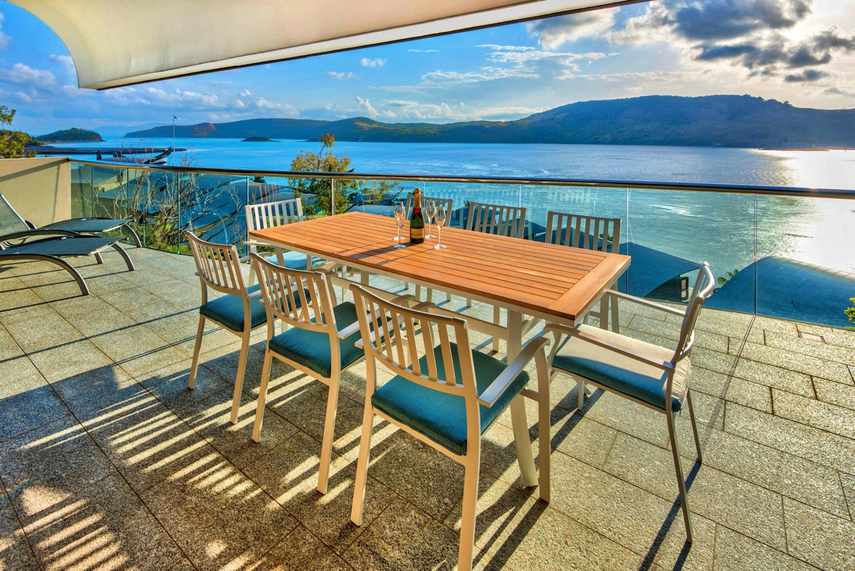 Une table en bois, six chaises, une bouteille de champagne et deux verres sont disposés sur un balcon donnant sur une étendue d’eau et sur des collines lointaines.