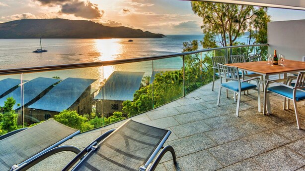 A balcony with a view of the sea, a table with chairs, and a bottle of wine. - Waves 5 Luxury 3 Bedroom (Whitsundays)
