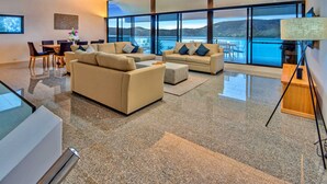 A spacious living room with a large window offering a view of the water and hills. There are two sofas and a dining table with chairs. - Waves 5 Luxury 3 Bedroom (Whitsundays)
