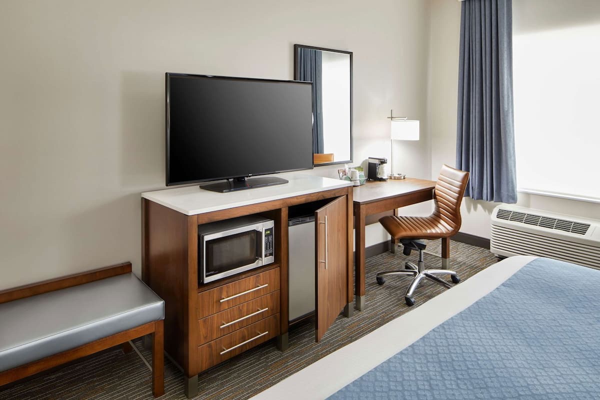 A hotel room with a bed, a desk with a television, a microwave, a chair, a bench, and a window with curtains.