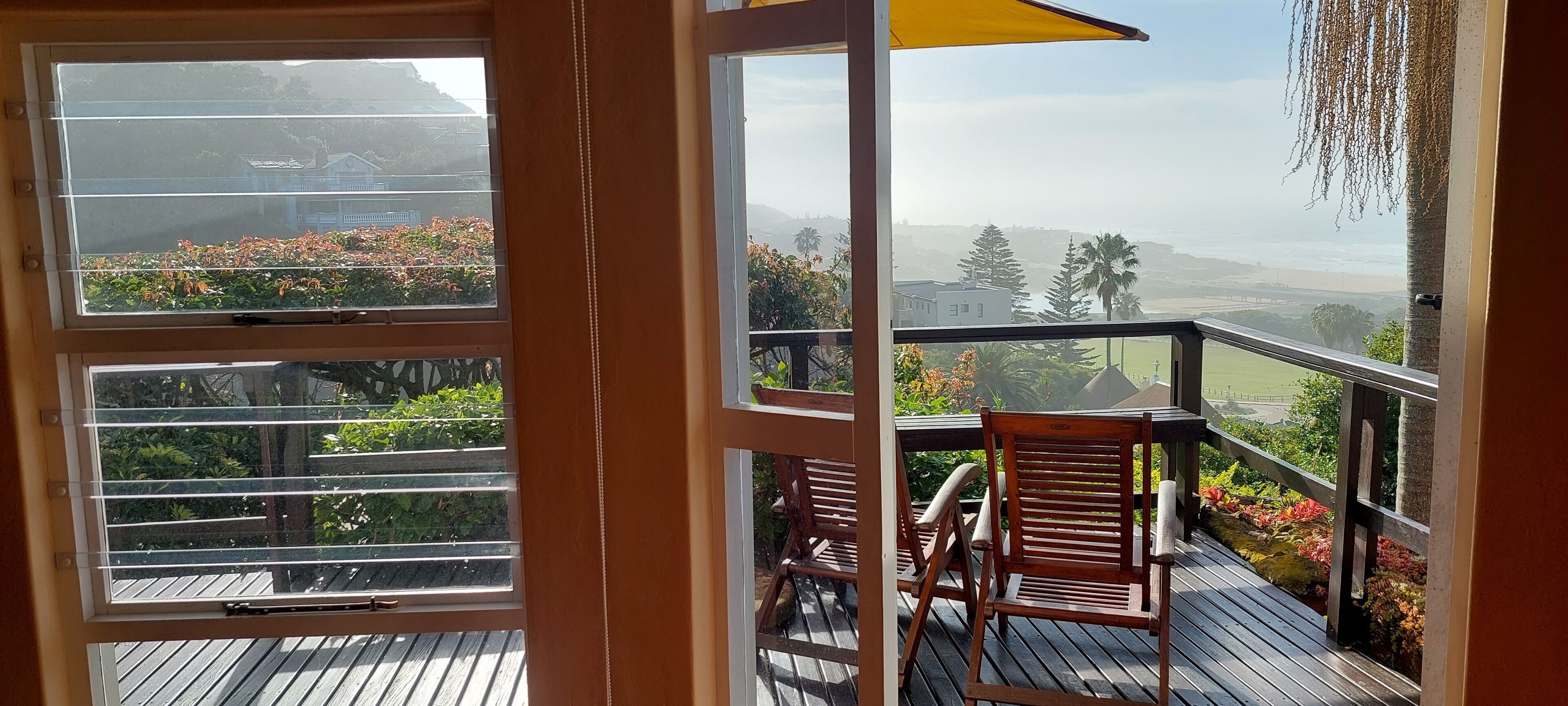 A wooden deck with a table and chairs, overlooking a garden with various plants and a distant view of a building and landscape.