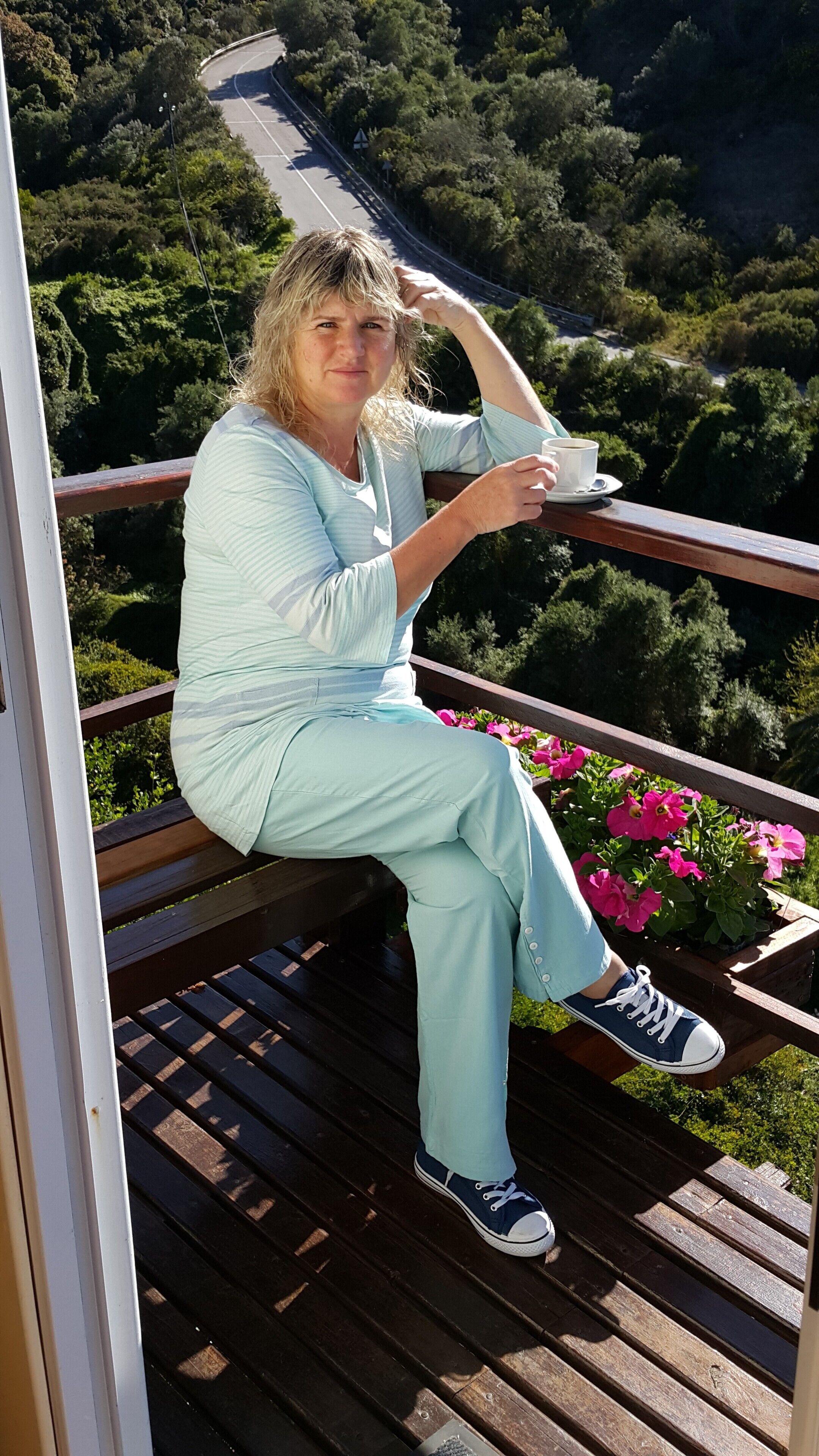 A person sitting on a wooden deck with a railing, overlooking a road and greenery.