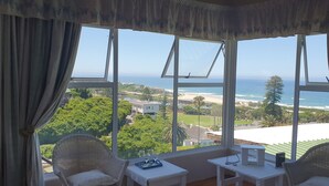 A room with a view of the ocean, a wicker chair, a small table with glasses and a tray, and a window with curtains. - The Tops (Wilderness)