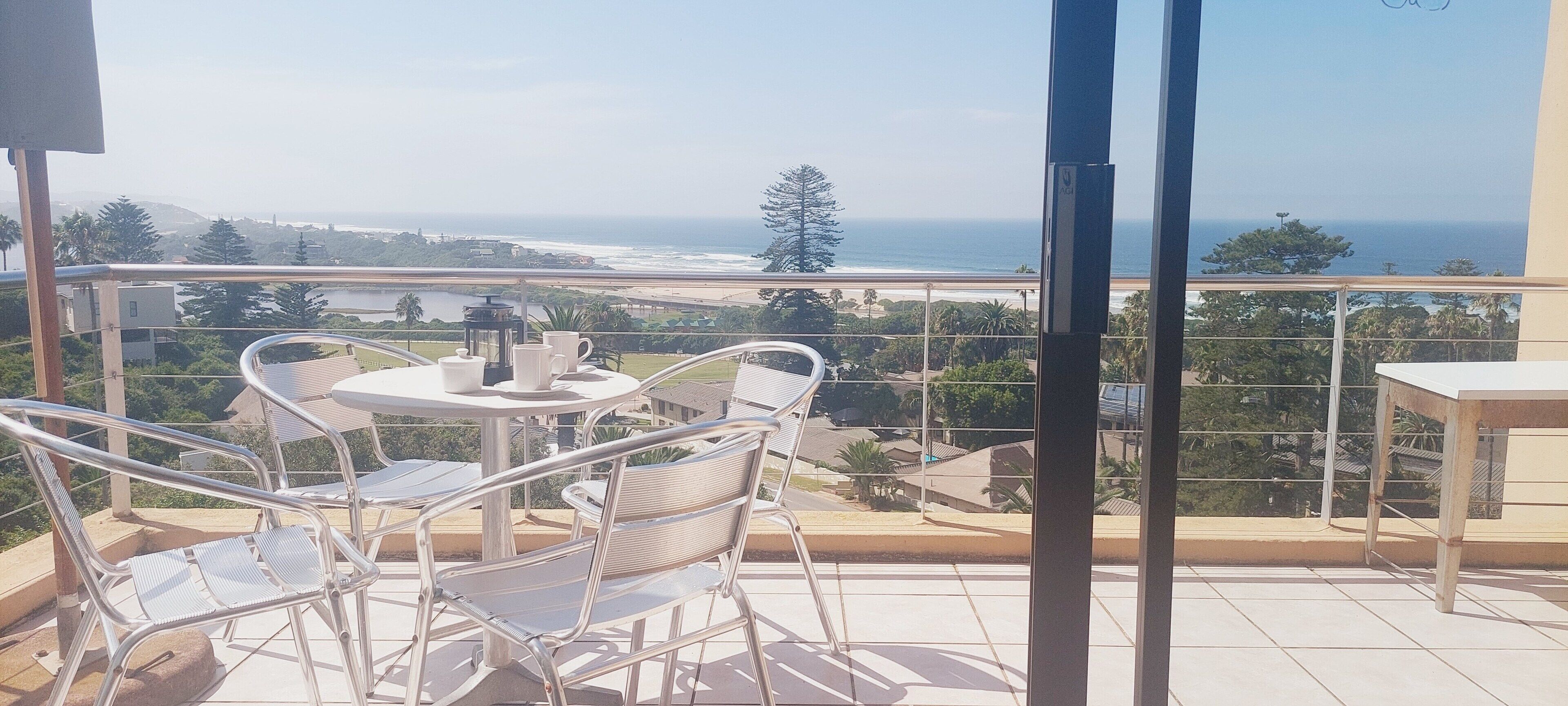 A balcony with a table and chairs, overlooking a coastal view with a beach and ocean.