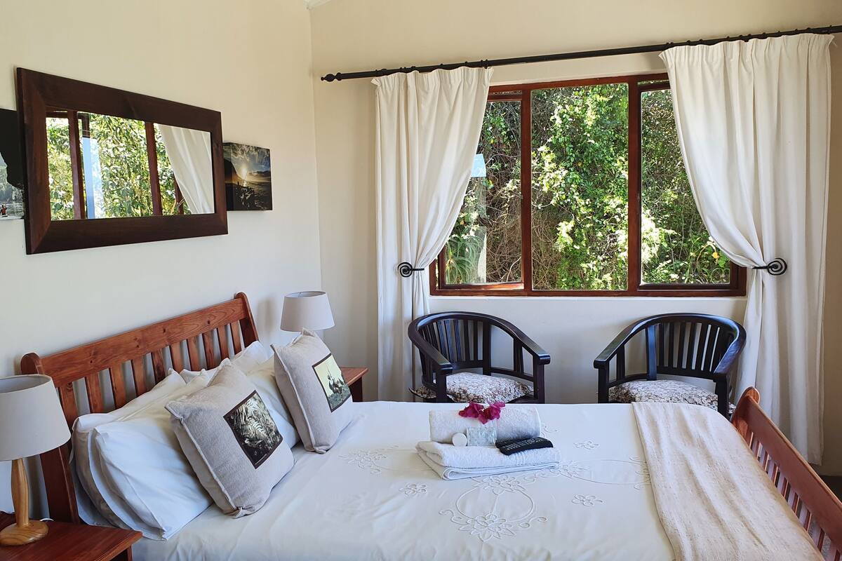 A bedroom with a wooden bed, two chairs, a nightstand with a lamp, and a window with curtains.