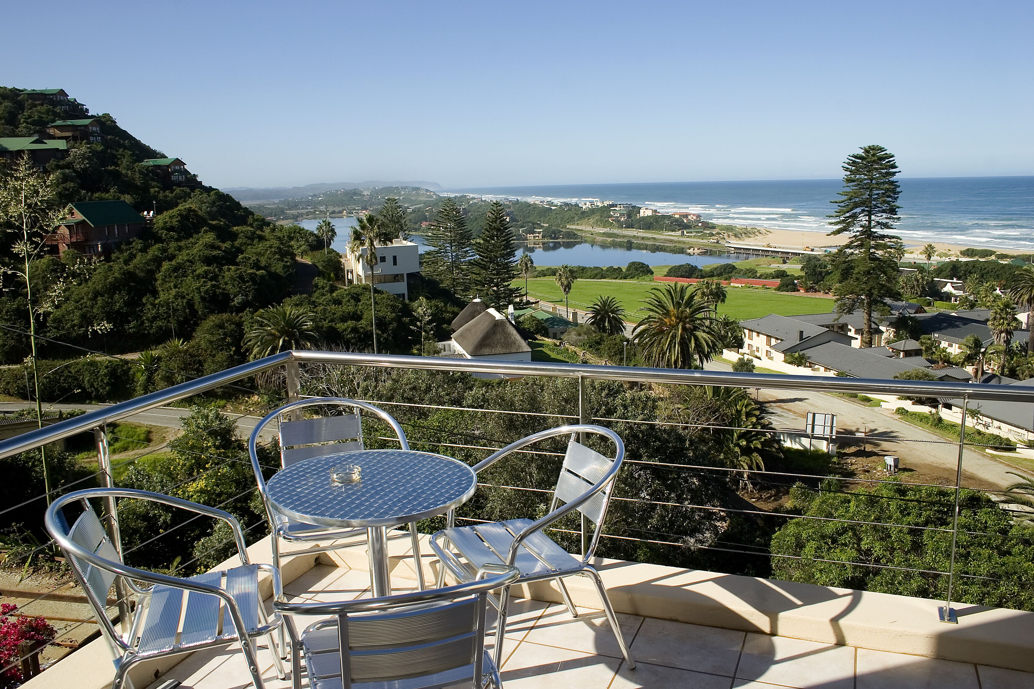 Un balcon avec une table et des chaises donnant sur un paysage côtier, avec des maisons, une plage et une étendue d’eau.