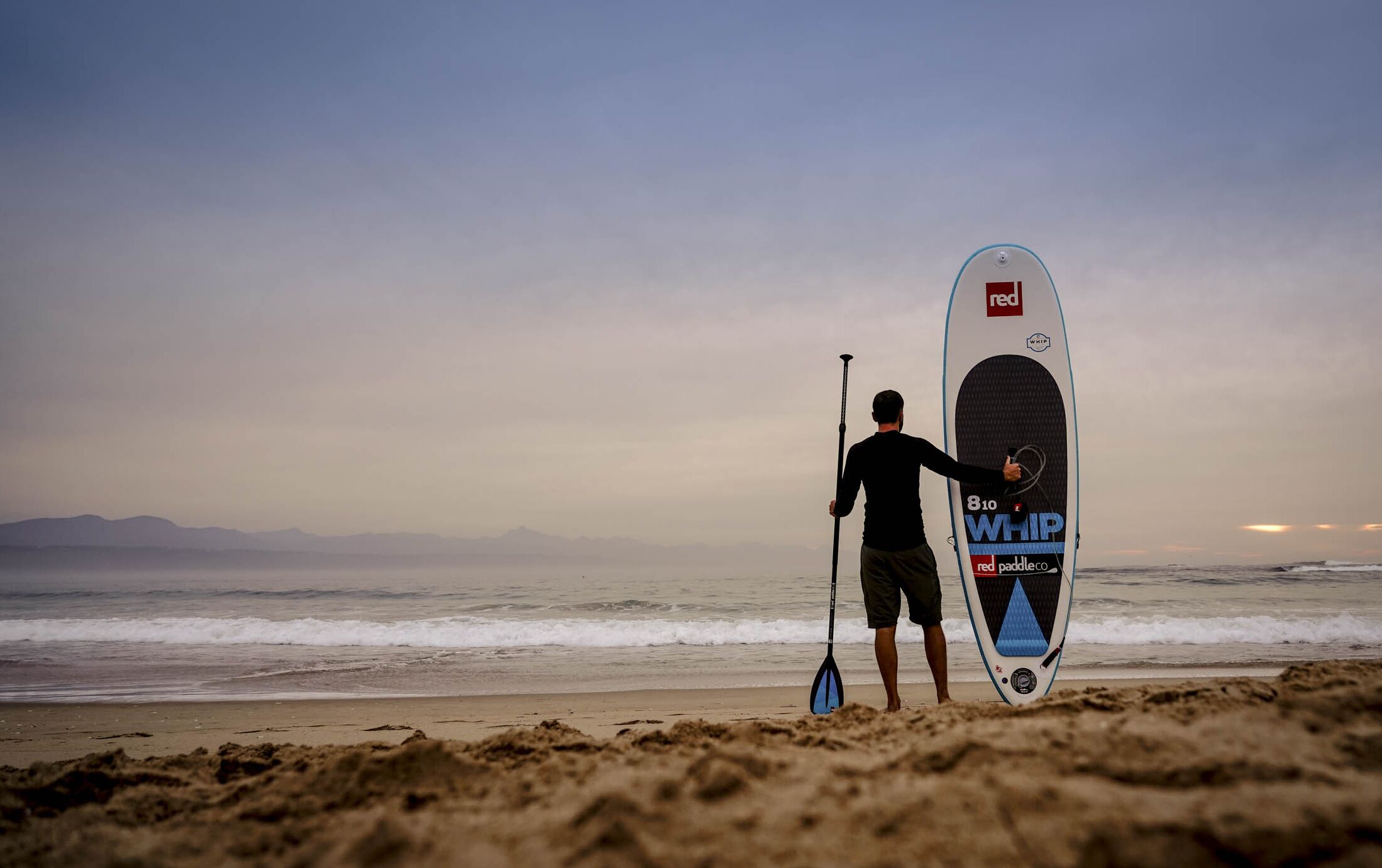 Una persona de pie en la playa sosteniendo una tabla de remo.