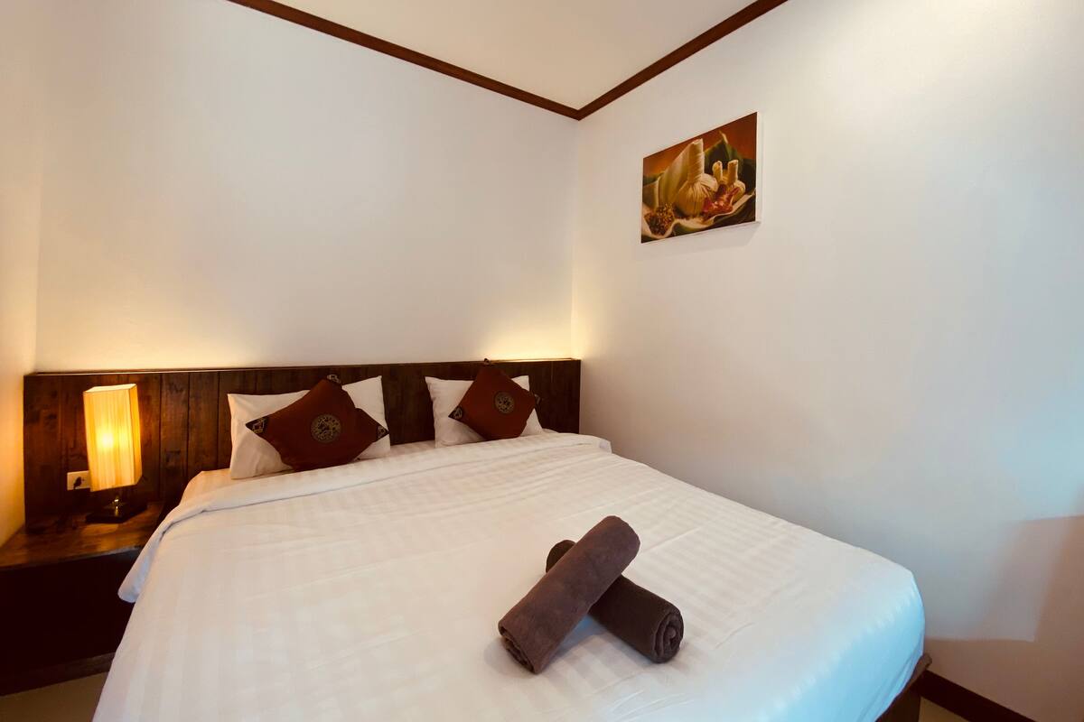 A hotel room with a neatly made bed, a headboard with a wooden panel, a nightstand with a lamp, and a painting on the wall.