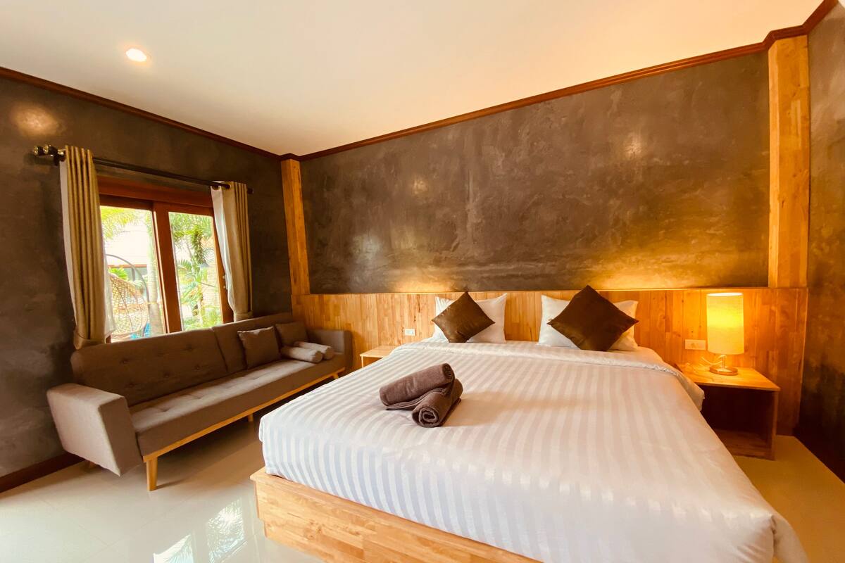 A modern bedroom with a large bed, a wooden headboard, a sofa, and a bedside table with a lamp.