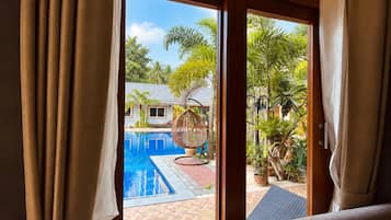 Blick von einem Zimmer mit großem Fenster auf einen Swimmingpool, ein Korbstuhl und ein Topfpflanze.