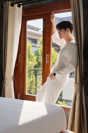 A person standing by a window with curtains, a bed with a white mattress, and a view of greenery outside. - Paradise Land Resort (Pattaya)