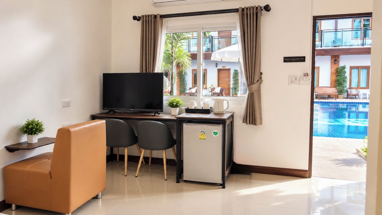 Quarto de hotel com televisão, duas cadeiras, mini-frigorífico, mesa de trabalho e vista para uma piscina.