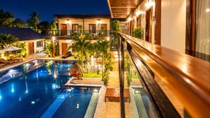 Una piscina con barandilla de vidrio, rodeada de sillas de descanso y plantas en macetas, con un edificio al fondo.