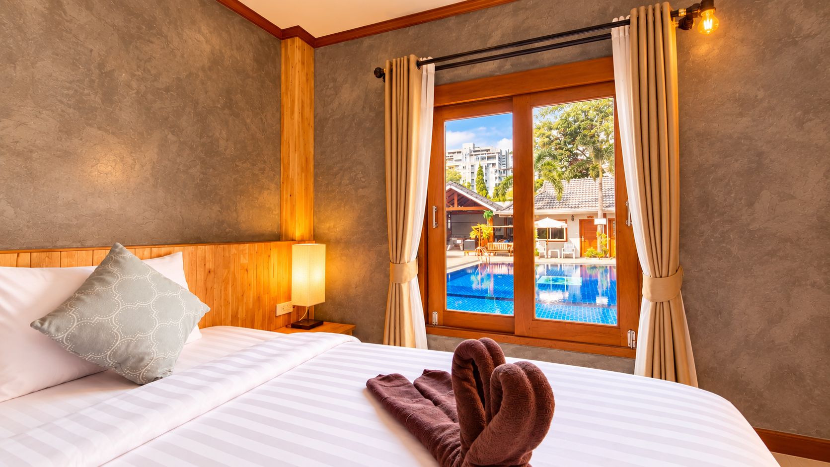 A hotel room with a bed, a nightstand with a lamp, a window with curtains, and a view of a pool and buildings outside.