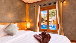 A hotel room with a bed, a nightstand with a lamp, a window with curtains, and a view of a pool and buildings outside. - Paradise Land Resort (Pattaya)
