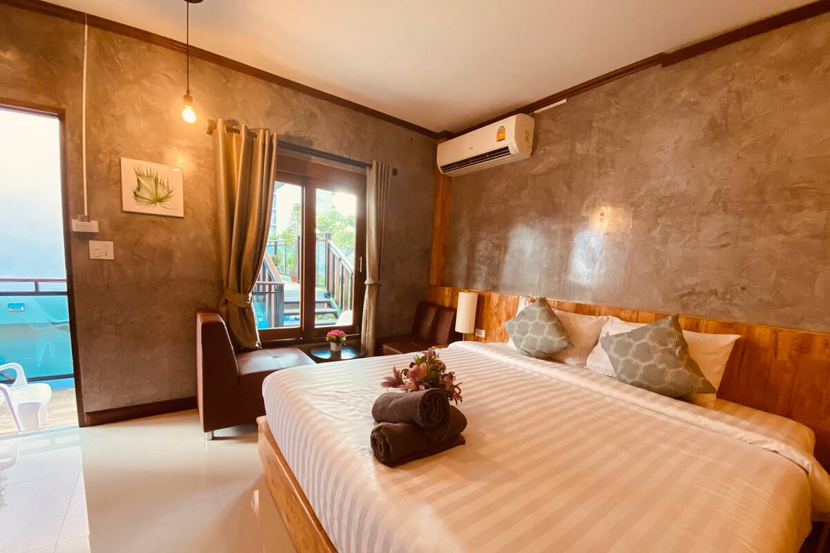 A hotel room with a large bed, a desk, a chair, a small table with a vase of flowers, and a view of a pool and deck area.