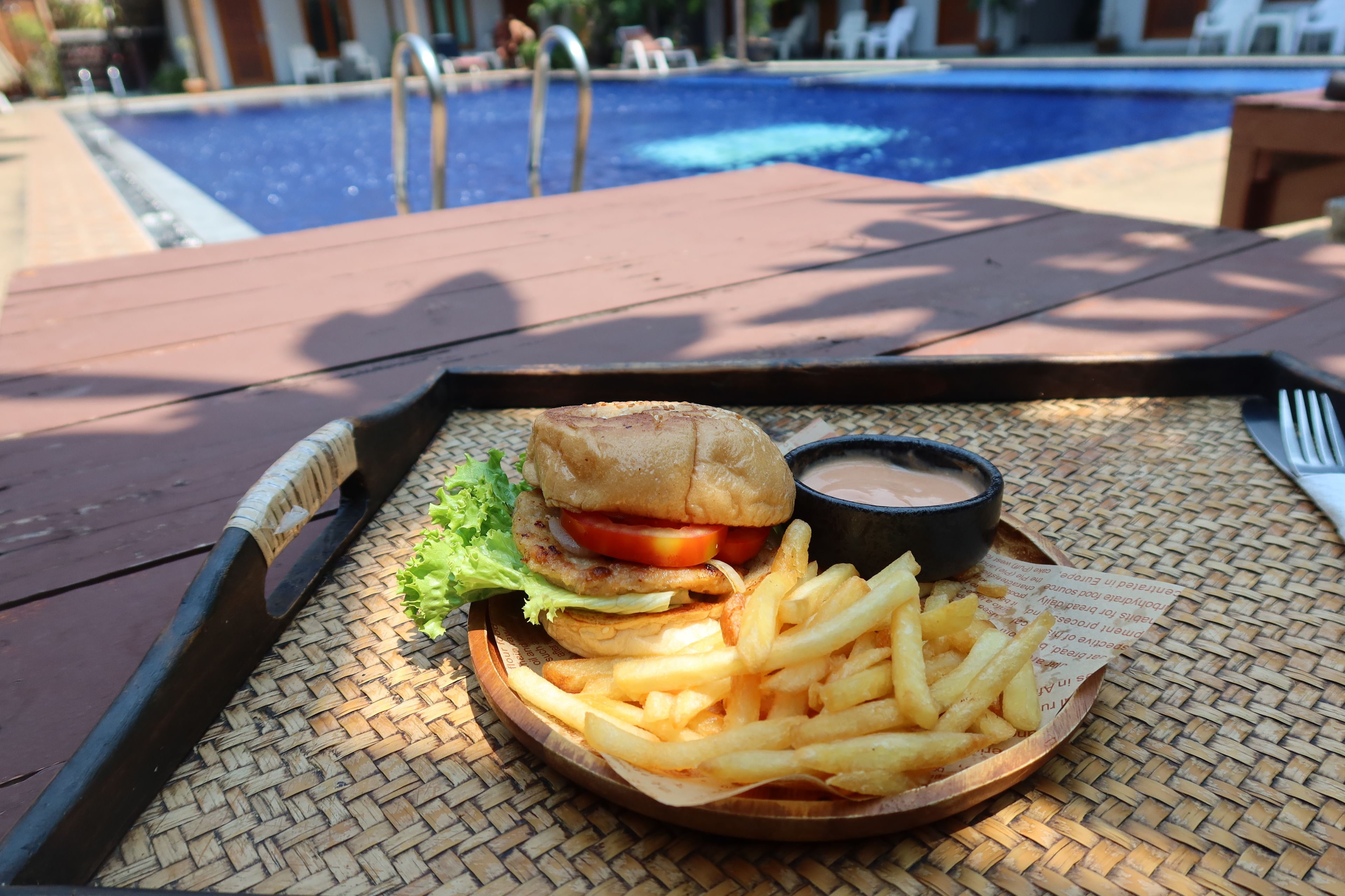 A meal consisting of a burger, fries, and a dipping sauce on a woven tray, placed on a table with a pool in the background.