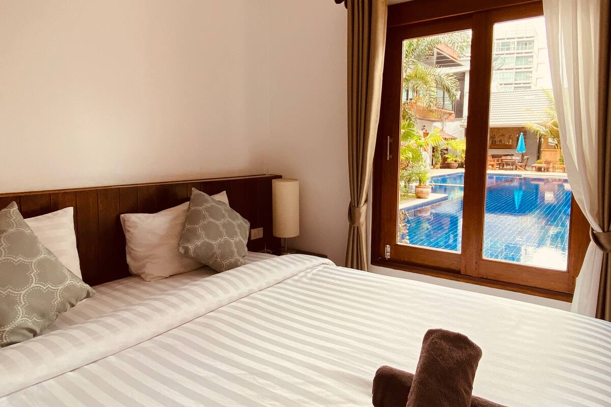 A hotel room with a bed, pillows, a nightstand, and a view of an outdoor swimming pool.
