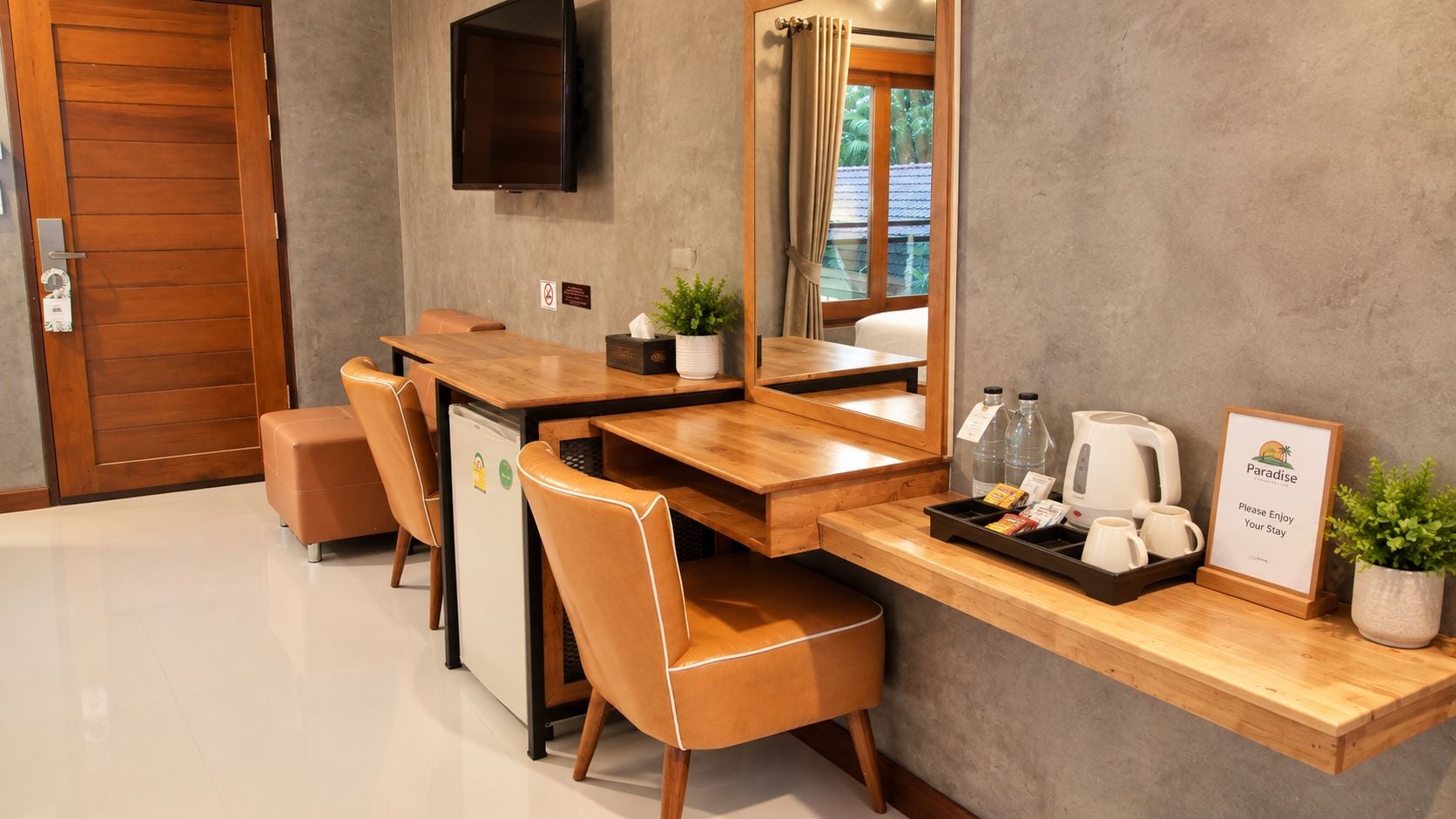 A hotel room with a wooden desk, chairs, a television, a small refrigerator, a coffee station, and a welcome sign.