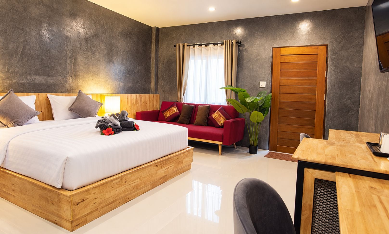 Ein modernes Hotelzimmer mit einem hölzernen Bett, einem roten Sofa, einem Esstisch und einer großen Pflanze.