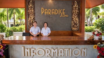 Un mostrador de recepción con letreros que dicen 'PARADISE LAND' e 'INFORMACIÓN', dos empleados y vistas a un jardín tropical.