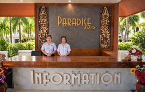 Un mostrador de recepción con letreros que dicen 'PARADISE LAND' e 'INFORMACIÓN', dos empleados y vistas a un jardín tropical.