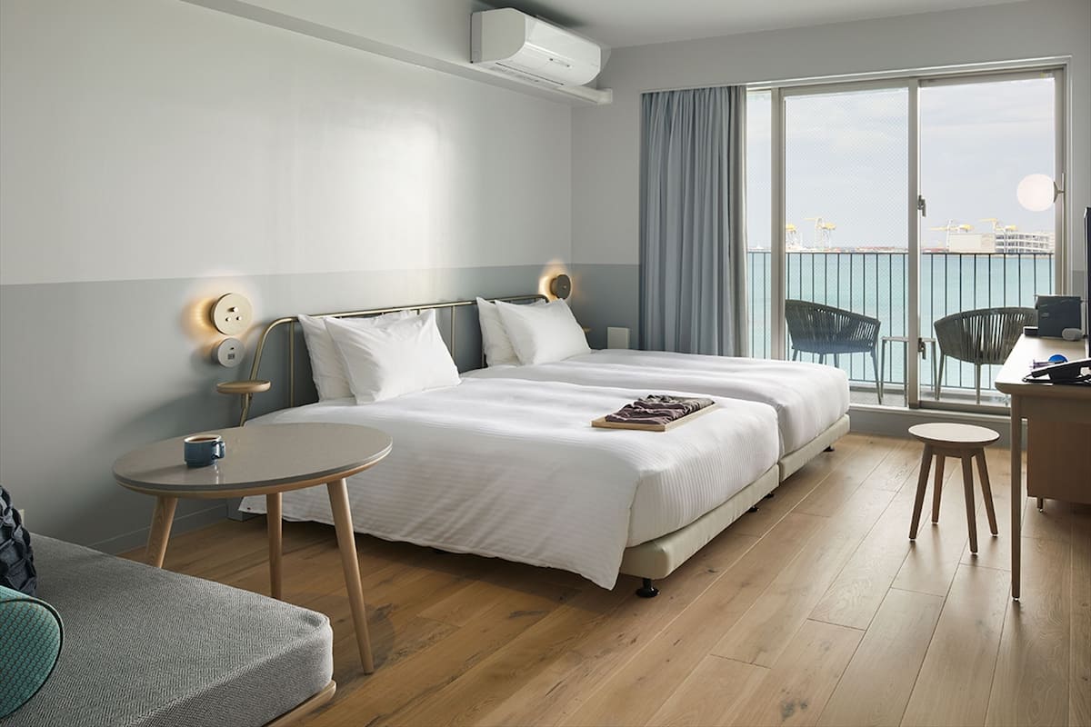 A modern hotel room with a large bed, a small round table, a desk, and a balcony with a view of the sea.