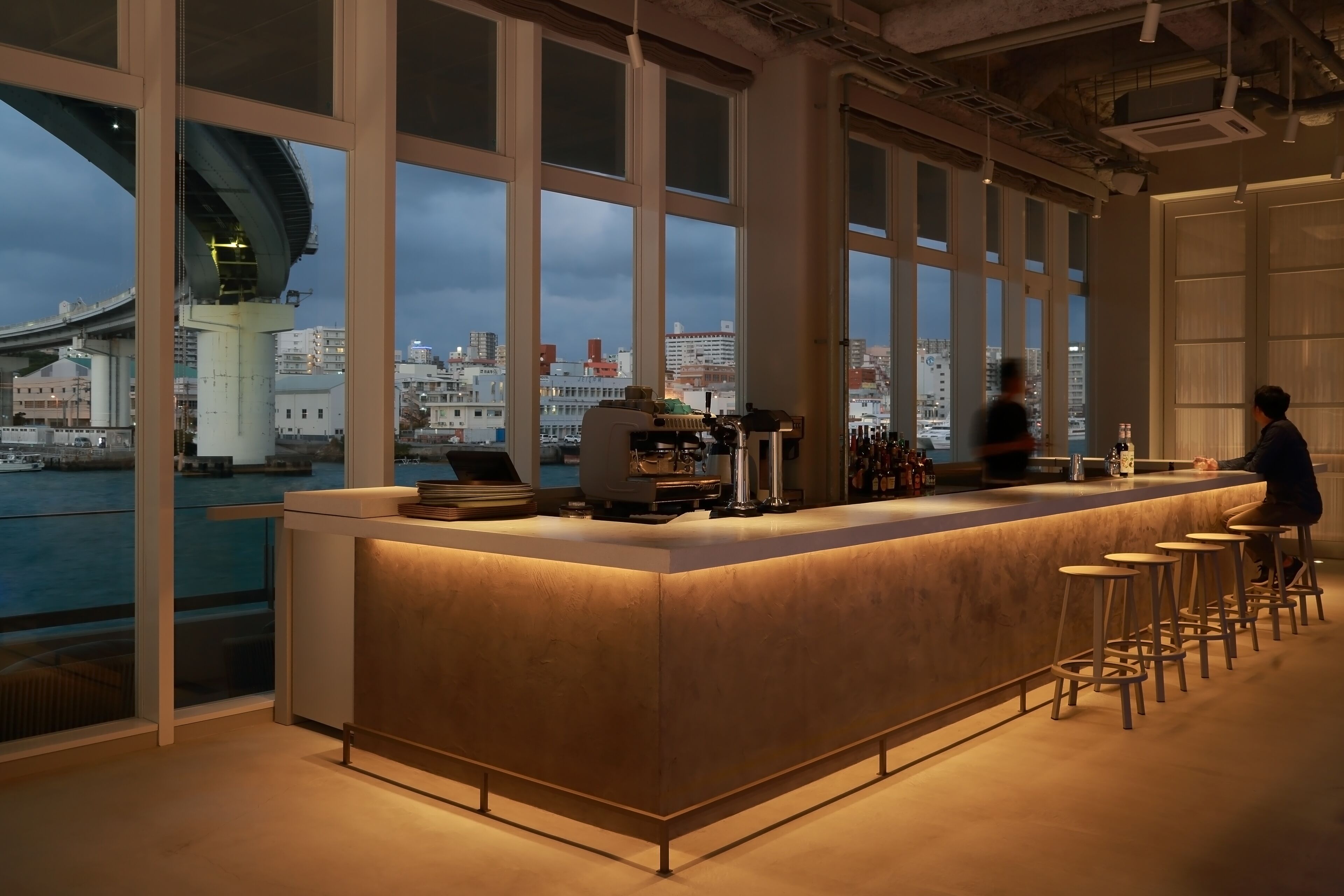 A bar with a long counter and stools, large windows, and a view of a bridge and cityscape.
