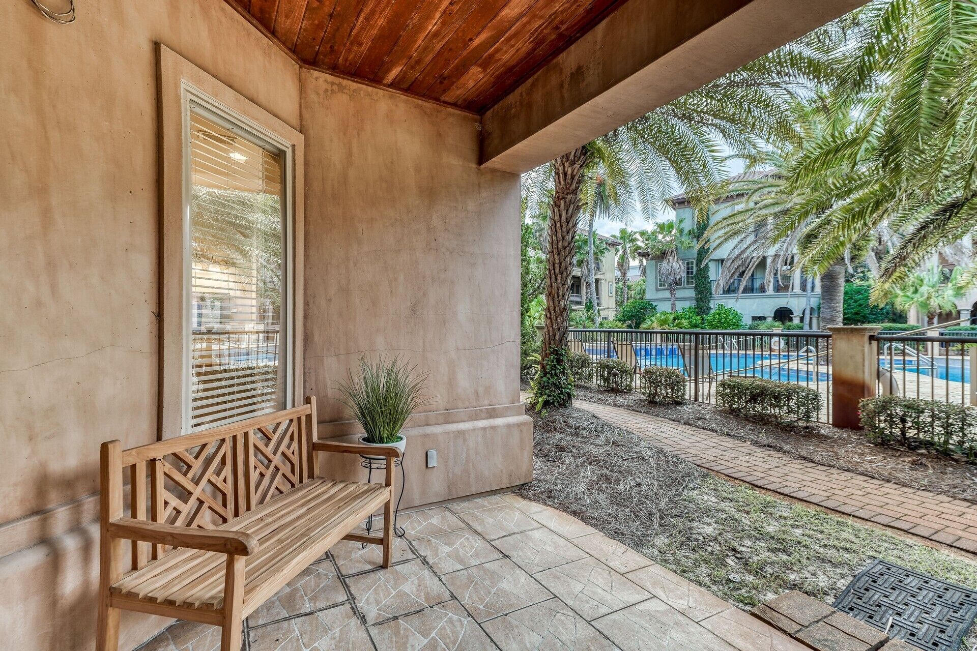 A covered outdoor area with a wooden bench, a potted plant, and a view of a swimming pool and palm trees.