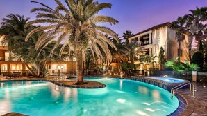 Ein Swimmingpool mit einer zentralen Palme, umgeben von einer Poolanlage mit Liegestühlen und einem Gebäude im Hintergrund.