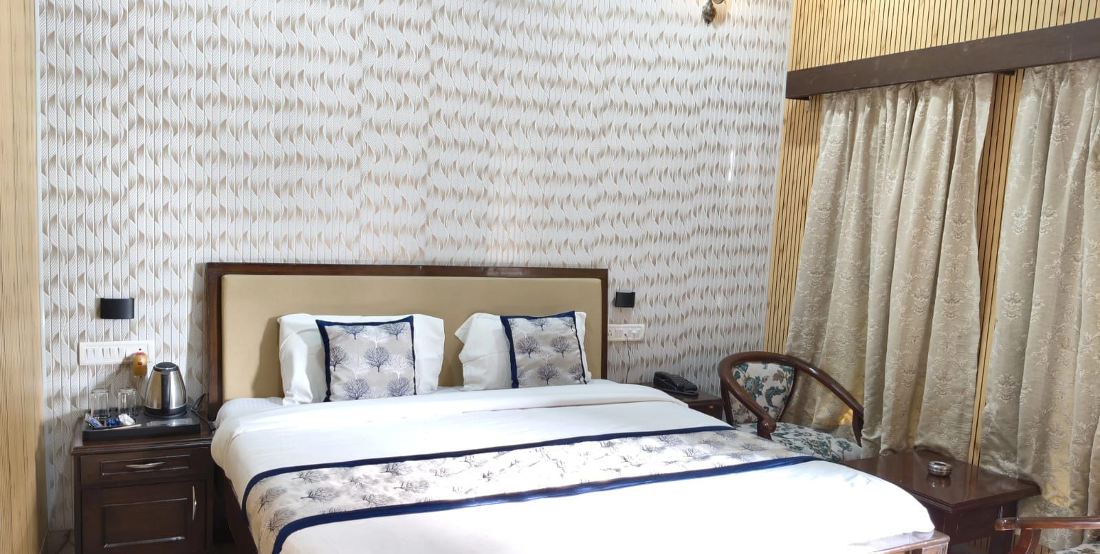 A hotel room with a bed, bedside tables, a chair, and a patterned wallpaper.