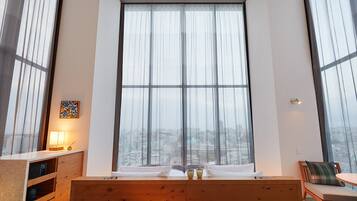 A modern hotel room with a large window, a wooden bed, a bedside table with a lamp, and a view of the cityscape.
