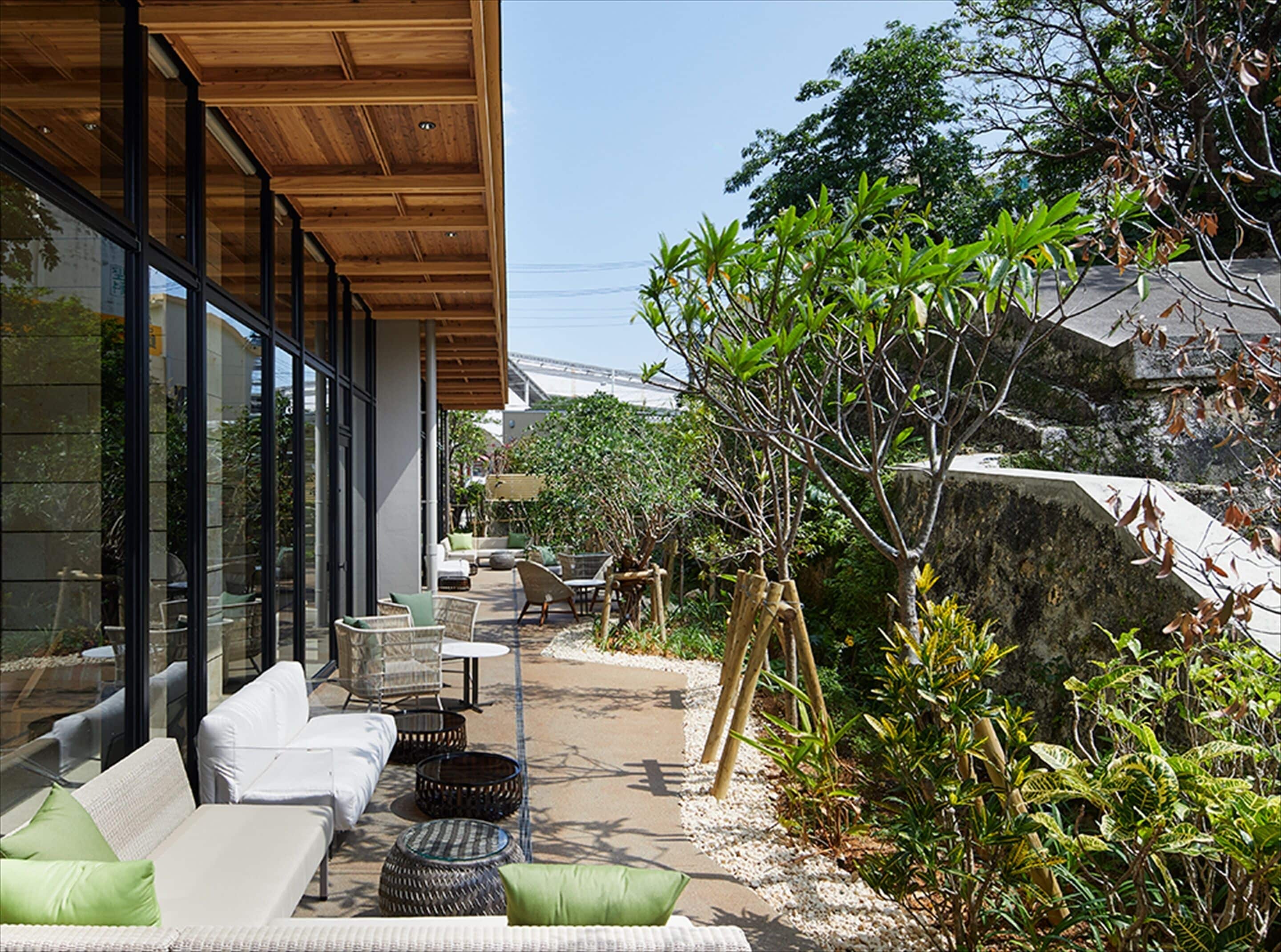 A modern outdoor seating area with white sofas and wicker chairs, surrounded by greenery and a stone wall.