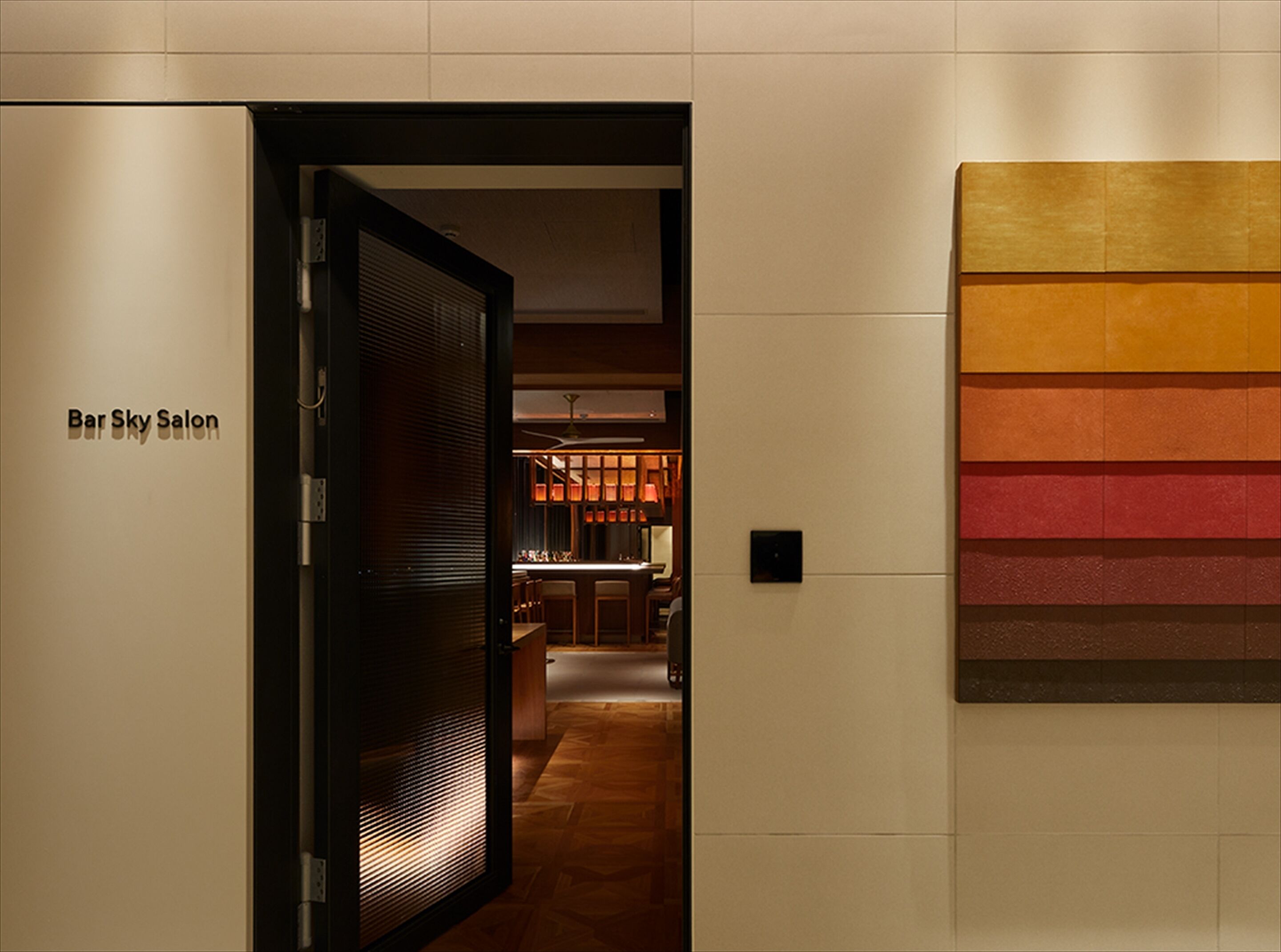 Un pasillo con una puerta que indica “Bar Sky Salon” que da a un área de bar con una decoración moderna y una pared con bandas horizontales de color.