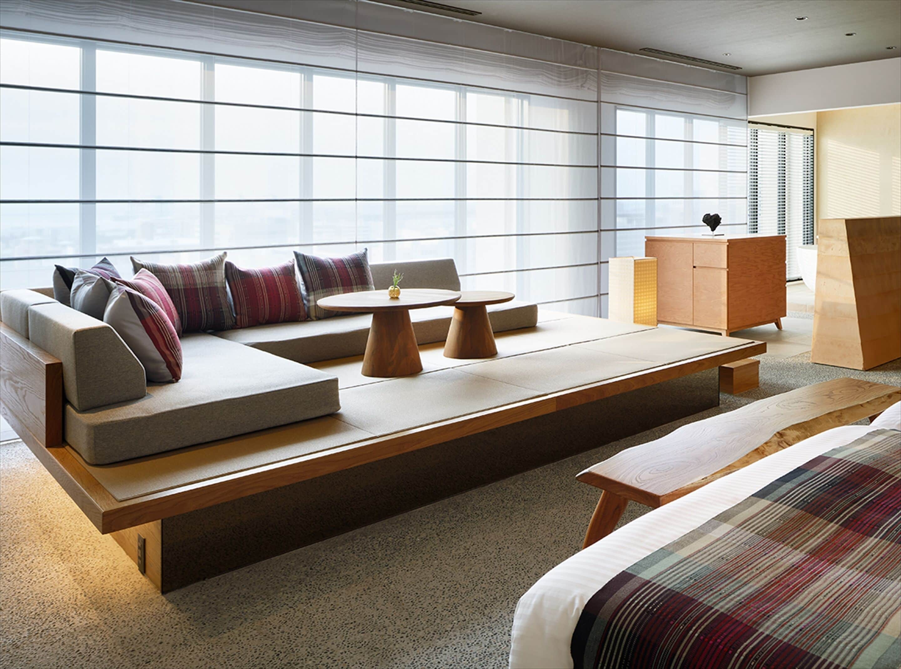 A modern hotel room with a large window, a sofa with multiple cushions, a wooden coffee table, a small side table, and a wooden cabinet.