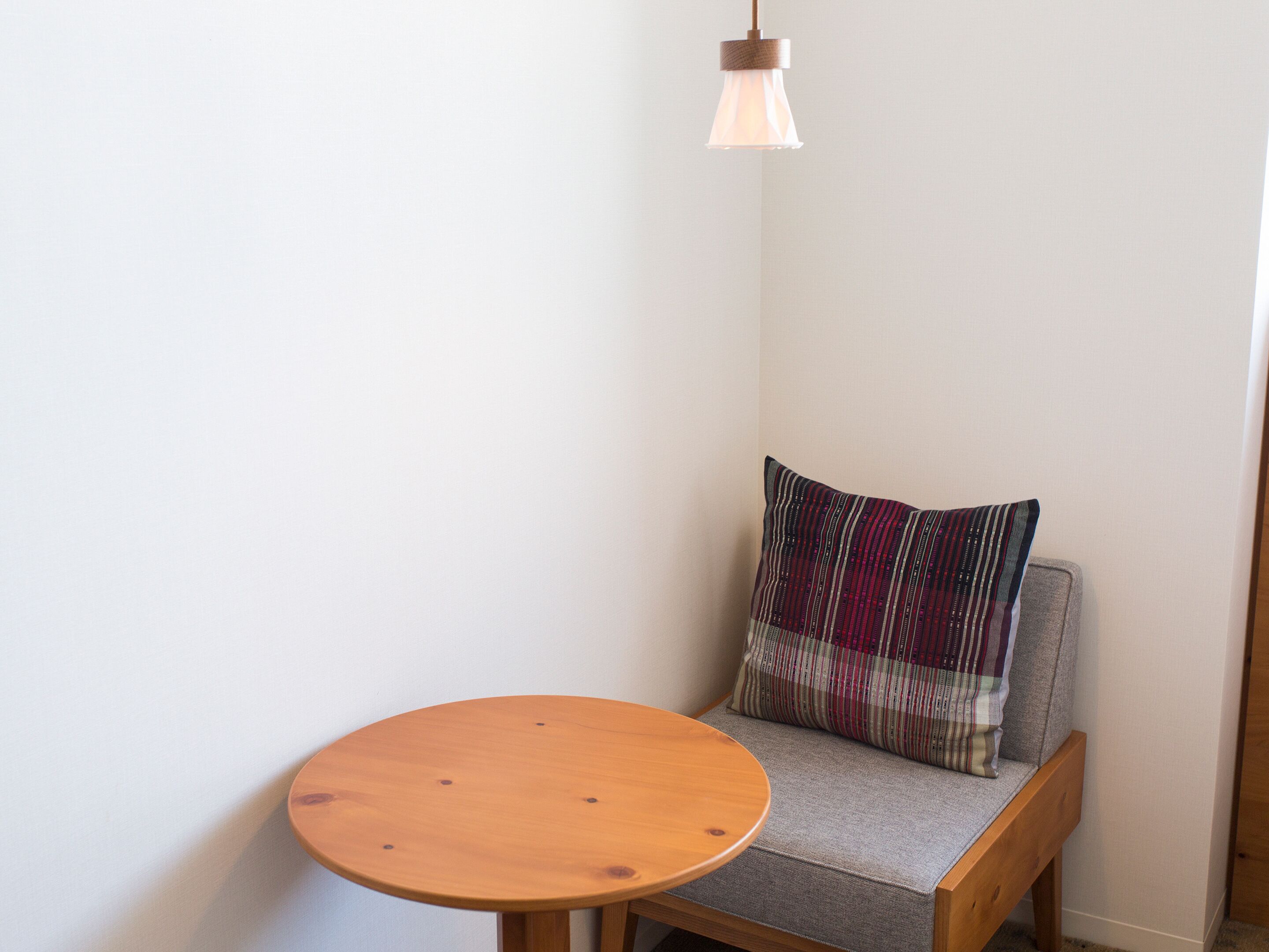 Un coin de pièce avec une table ronde en bois, un banc rembourré agrémenté d’un coussin à motifs et un luminaire suspendu.