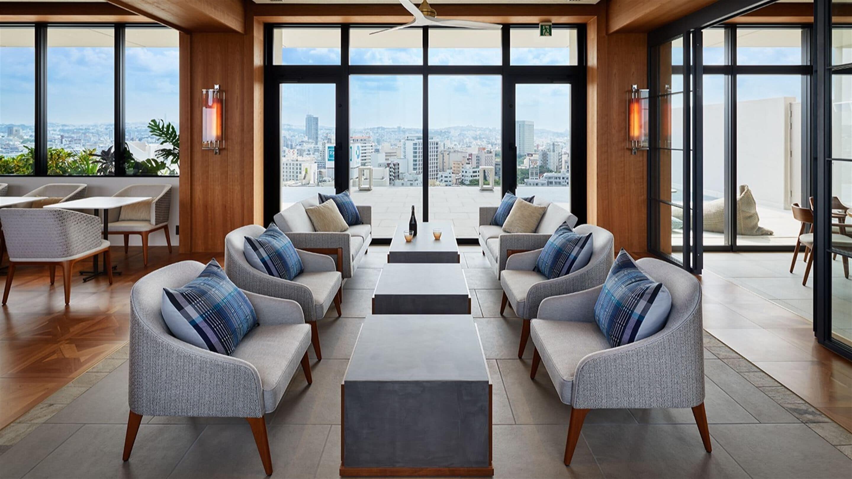 A modern lounge area with sofas and tables, large windows offering city views, and a wooden ceiling.